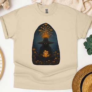 Może przedstawiać: Beżowa koszulka z ciemną ilustracją postaci w kapeluszu czarownicy otoczonej świecącymi grzybami. Postać trzyma grupę grzybów, a z kapelusza wydobywa się więcej grzybów i świecące światło.