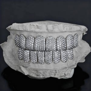 May include: A set of silver-colored grillz, encrusted with numerous small, sparkling stones, displayed on a white dental mold. The grillz are designed to fit over the upper and lower teeth, showcasing a luxurious aesthetic.
