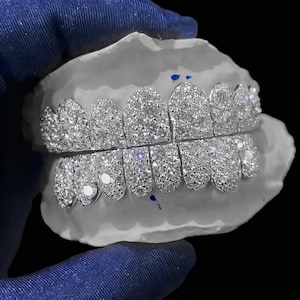 Puede incluir: Un juego de grillz plateados, incrustados con numerosas piedras pequeñas y brillantes. Los grillz se exhiben en un molde dental blanco, mostrando el intrincado diseño y la artesanía. El fondo es negro.