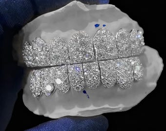 May include: A set of custom-fitted, silver-colored grillz encrusted with numerous small, sparkling stones. The grillz are displayed on a white dental mold, showcasing the intricate design and craftsmanship. The background is black.
