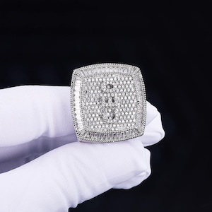 May include: A square, silver ring with a large, sparkling surface. The ring is encrusted with numerous small, clear stones, with a border of rectangular stones. The ring is held by a gloved hand against a black background.