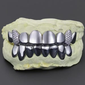 Può includere: Grillz color argento con finitura lucida, posizionati su un calco dentale giallo chiaro. I grillz presentano una fila di denti superiori e inferiori, con accenti di diamanti sui denti laterali. Lo sfondo è grigio.