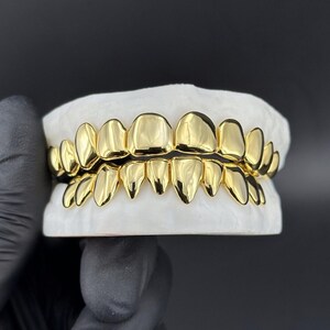 May include: A set of gold-colored teeth grillz, displayed on a white mold. The grillz are highly polished, reflecting light, and feature a full set of upper and lower teeth. The background is a solid black.