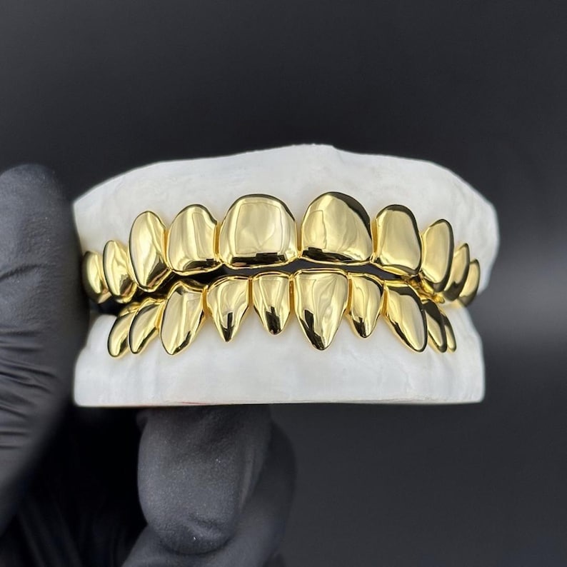 May include: A set of gold-colored teeth grillz, displayed on a white mold. The grillz are highly polished, reflecting light, and feature a full set of upper and lower teeth. The background is a solid black.