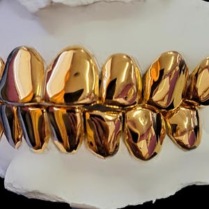 May include: Close-up of a set of gold teeth. The teeth are highly polished, reflecting light and appearing bright gold. The gold teeth are set on a white base, creating a striking contrast. The image showcases dental jewelry.