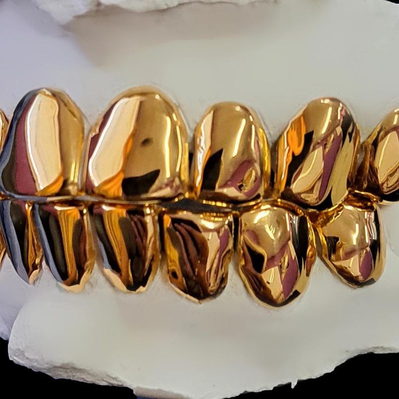 May include: Close-up of a set of gold teeth. The teeth are highly polished, reflecting light and appearing bright gold. The gold teeth are set on a white base, creating a striking contrast. The image showcases dental jewelry.