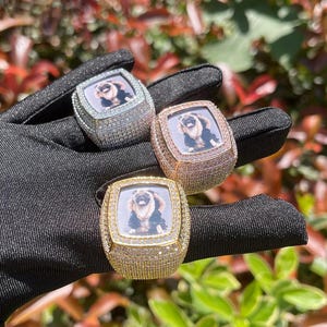 May include: Three square-shaped rings with a photo of a dog inside. The rings are set with clear, pink, and yellow stones.