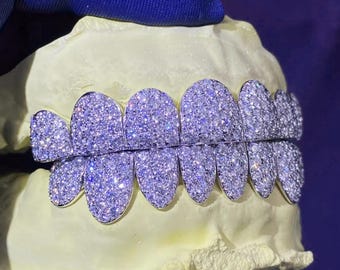 May include: A set of custom-made, diamond-encrusted grillz. The grillz are silver-toned and fully covered in small, sparkling diamonds. They are displayed on a light yellow dental mold against a blue background.