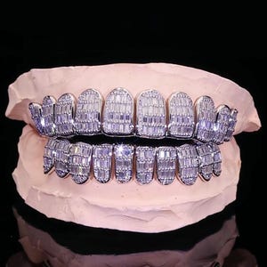 Puede incluir: Un juego de grillz plateados hechos a medida con gemas claras de corte rectangular. Los grillz se exhiben sobre un molde dental rosa sobre un fondo negro. Se ven las filas de dientes superiores e inferiores.