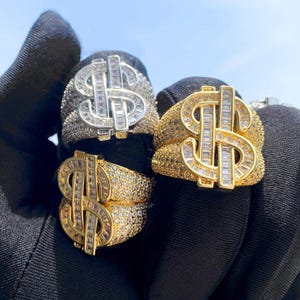 May include: Three gold and silver rings featuring a dollar sign design. The rings are set with clear cubic zirconia stones.