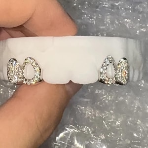 May include: A white dental mold with two sets of silver-colored, gem-studded tooth caps. The caps are designed to fit over the front teeth, with a sparkling, decorative appearance. The gems are clear and reflect light.