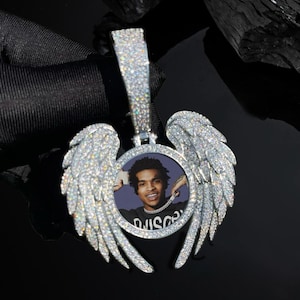 May include: A silver pendant featuring a photo of a person, framed by diamond-set angel wings. The pendant has a rectangular bail. The background is black.