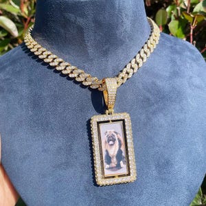 May include: Gold-toned necklace with a rectangular pendant featuring a photo of a pug. The pendant and chain are encrusted with clear, sparkling stones. The necklace is displayed on a blue surface.