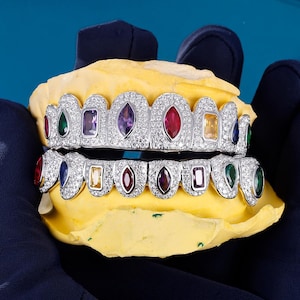 May include: A set of custom-made silver grillz with colorful gemstones. The grillz are set with a variety of gems, including red, green, purple, and yellow stones. The grillz are displayed on a yellow dental mold against a blue background.