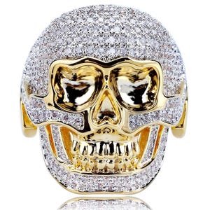 May include: A gold-toned skull ring with a detailed design. The skull's cranium is encrusted with small, clear gemstones, while the face features a polished gold finish. The ring is a bold statement piece.