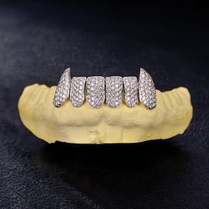 May include: A set of silver-colored, diamond-studded teeth grillz, displayed on a yellow dental mold. The grillz feature a row of six teeth, with the two outer teeth designed as fangs. The diamonds sparkle against the dark background.