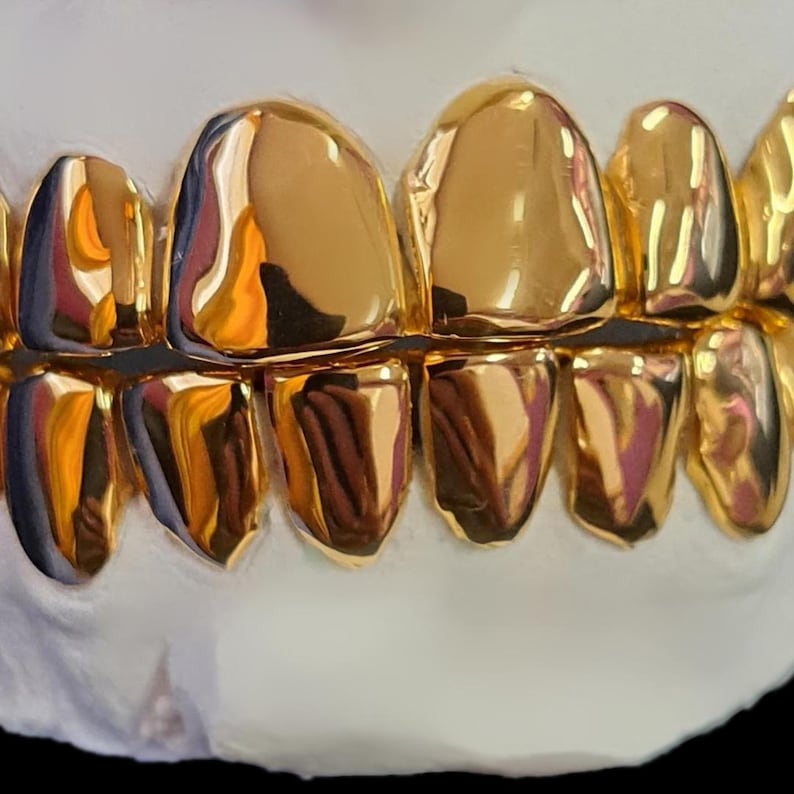 May include: Close-up of a set of gold teeth, showcasing a gleaming, polished surface. The teeth are a bright, reflective gold color, with a smooth, rounded shape. The image highlights the dental work's craftsmanship and luxurious appearance.