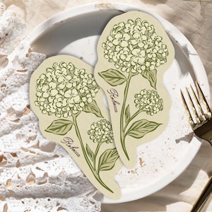 Może przedstawiać: Dwie wycinane naklejki z botanicznymi ilustracjami kwiatów hortensji w odcieniach jasnej zieleni i kremu. Każda naklejka przedstawia szczegółowy kwiatostan z liśćmi oraz kursywny napis Rebecca wydrukowany wzdłuż łodygi.