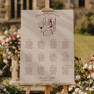 May include: A cream-coloured wedding seating plan displayed on a wooden easel. The chart has a decorative illustration and the text "Please find your seat." Names are listed under numbers one to twelve. The names "Lauren & Jack" and the date "20th June 2021" are also printed on the chart.