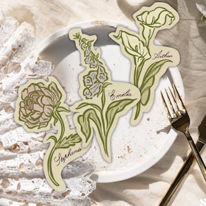 Könnte beinhalten: Drei gestanzte Platzkarten aus Papier mit botanischen Illustrationen einer Pfingstrose, eines Fingerhuts und einer Calla-Lilie in Grün- und zarten Rosatönen. Jede Karte zeigt einen handgeschriebenen Namen: Stephanie, Emilia und Arthur in Dunkelbraun.