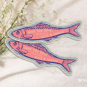 May include: Two fish-shaped cards featuring a pink fish illustration with blue outlines and blue striped background. The text reads SAVE THE DATE 20.06.2023 ALICE & MARCUS. The cards are positioned next to a bouquet of dried flowers with a white ribbon.