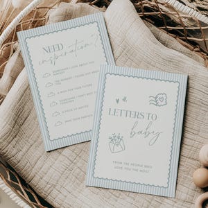 May include: Two baby shower cards with blue striped borders and text, one reading "Need Inspiration?" and the other "Letters to Baby." The cards are on a beige cloth inside a white woven basket, with wooden toys and dried plants.
