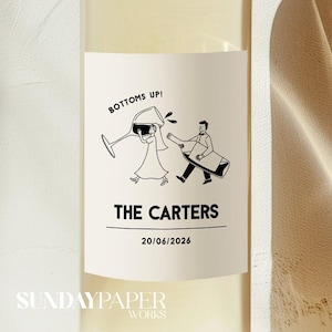 Pode incluir: Uma garrafa de vinho com um rótulo com uma ilustração a preto e branco de um casal e uma taça de vinho. O texto no rótulo diz "BOTTOMS UP!", "THE CARTERS" e uma data.