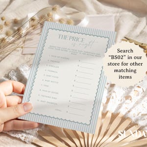 May include: A baby shower game card with a light blue striped border and the text "The Price is Right". The card includes a list of baby essentials. The image also includes a pair of gold scissors and a fan.