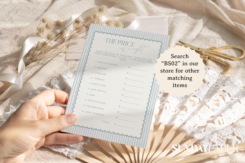 May include: A baby shower game card with a light blue striped border and the text "The Price is Right". The card includes a list of baby essentials. The image also includes a pair of gold scissors and a fan.