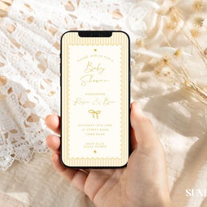 Może przedstawiać: Smartfon wyświetla zaproszenie na baby shower w kremowo-złotej kolorystyce. Tekst brzmi "Please join us for a Baby Shower Honoring Rose & Ben." Telefon jest trzymany w dłoni, z koronkowym wachlarzem i suszonymi kwiatami w tle.