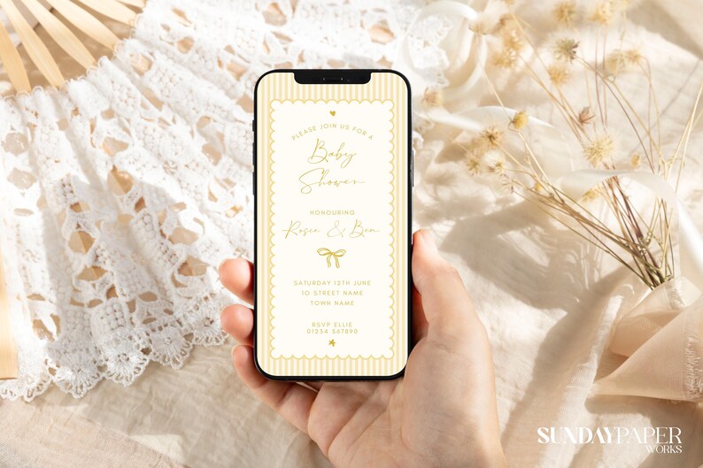 Może przedstawiać: Smartfon wyświetla zaproszenie na baby shower w kremowo-złotej kolorystyce. Tekst brzmi "Please join us for a Baby Shower Honoring Rose & Ben." Telefon jest trzymany w dłoni, z koronkowym wachlarzem i suszonymi kwiatami w tle.