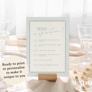 May include: A table sign with the text "Need Inspiration?" and prompts for reflection. The sign is light blue and white, with a scalloped border, and is held by a wooden base. Two clear glasses are on either side.