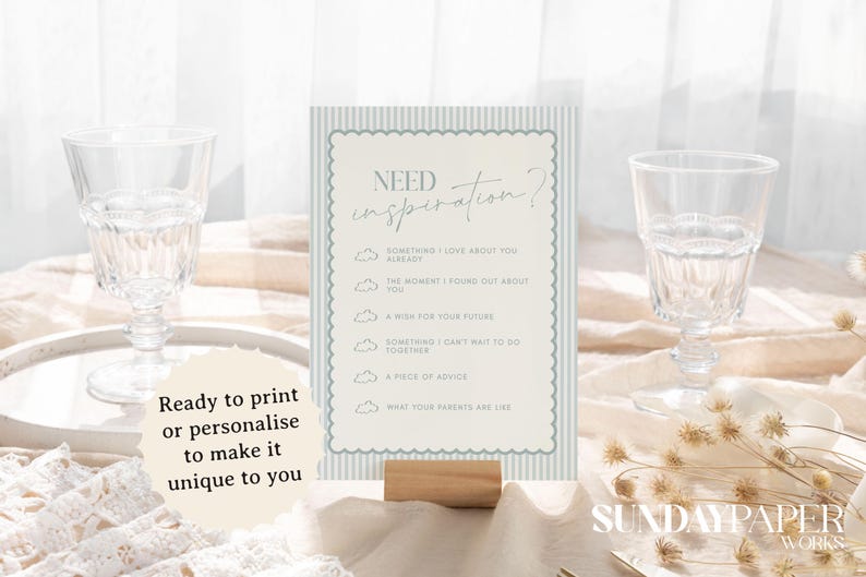 May include: A table sign with the text "Need Inspiration?" and prompts for reflection. The sign is light blue and white, with a scalloped border, and is held by a wooden base. Two clear glasses are on either side.