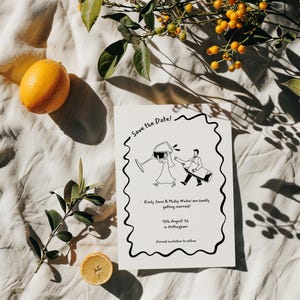 A fun, hand drawn wedding save the date card lies on a neutral linen tablecloth, with sliced and whole lemons and green leafy stems for decoration, giving the essence of an Italian or Spanish tablescape. The design of the card is fun, with a comical cartoon drawing of a bride and groom carrying a comically large wine glass and bottle