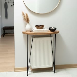 Può includere: Uno specchio rotondo con cornice dorata è appeso sopra un tavolino in legno con gambe in metallo nero. Il tavolo ospita un vaso bianco con piante secche, libri e due piccole ciotole di legno. Sotto c'è un termosifone.