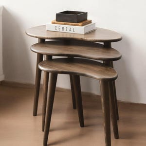 May include: A set of three nesting tables with a brown wood finish. The tables are shaped like a crescent moon and have three legs each. The top table has a book and a black box on it.
