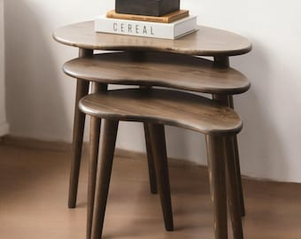 May include: A set of three nesting tables with a brown wood finish. The tables are shaped like a crescent moon and have three legs each. The top table has a book and a black box on it.