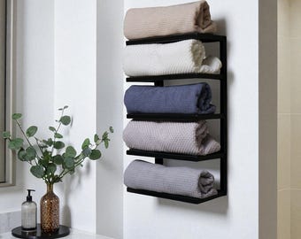 May include: A black metal towel rack mounted on a white wall, holding five rolled towels. The towels are in shades of beige, white, blue, gray, and light gray. The rack is in a bathroom setting.