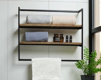 May include: A modern bathroom shelf with a black metal frame and wooden shelves. The shelves hold rolled towels in gray, beige, and blue, along with a jar and small bottles. A white towel hangs below. A potted plant sits nearby.