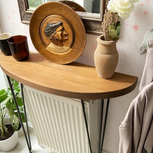 May include: A half-circle wooden console table with black hairpin legs. A decorative wooden plate with a carved profile is on the table, along with a small vase with flowers and two drinking glasses. A mirror hangs on the wall.