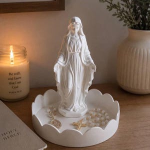 May include: A white matte statue of the Virgin Mary standing on a white scalloped tray. The tray holds a rosary with white beads and a metal crucifix. The statue features draped clothing and a head covering, with hands extended outward in a gesture.