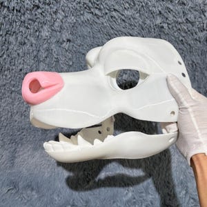 Könnte beinhalten: Weiße, unfertige Felltiermaske mit rosa Nase und offenem Mund. Die Maske besteht aus glattem, hartem Kunststoff und hat Löcher für Augen und Ohren.