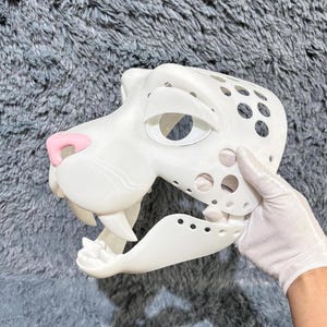 May include: White unfinished animal mask with a pink nose and open mouth. The mask has a detailed design with holes throughout.