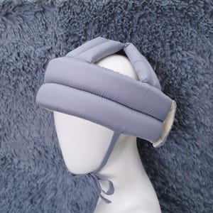 May include: A light blue padded baby safety helmet. The helmet is designed with a soft, quilted texture and features a chin strap for secure wear. The interior lining is a cream color.