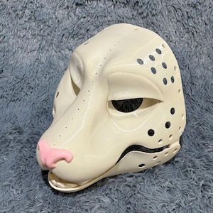 May include: A white, unpainted, plastic animal mask with a pink nose and holes for eyes and ears. The mask is designed to look like a dog or wolf.