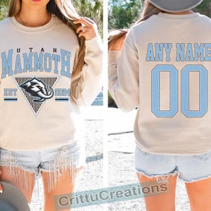 Puede incluir: Sudadera beige con "UTAH MAMMOTH" en letras azul claro y un logotipo de mamut. La parte trasera presenta "ANY NAME" y el n&uacute;mero "00" en azul claro. Una prenda c&oacute;moda para los aficionados al deporte.