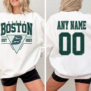 May include: White sweatshirt with "BOSTON" in green block letters, a logo, and "EST 2023". The back features "ANY NAME" above a green number "00". The sweatshirt is a casual style.