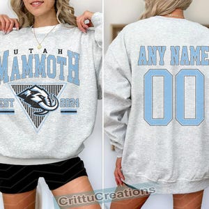 Puede incluir: Sudadera gris claro con "UTAH MAMMOTH" en letras azul claro. La parte delantera presenta un gr&aacute;fico triangular con un dise&ntilde;o de mamut y "EST 2024". La parte trasera muestra "ANY NAME" sobre el n&uacute;mero "00" en azul claro.