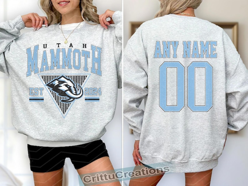 Puede incluir: Sudadera gris claro con "UTAH MAMMOTH" en letras azul claro. La parte delantera presenta un gr&aacute;fico triangular con un dise&ntilde;o de mamut y "EST 2024". La parte trasera muestra "ANY NAME" sobre el n&uacute;mero "00" en azul claro.