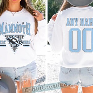 Puede incluir: Sudadera blanca con "UTAH MAMMOTH" en letras de bloque azul claro. La parte delantera presenta un logotipo del equipo y "EST 2024". La parte trasera muestra "ANY NAME" y el n&uacute;mero "00" en azul claro. Una prenda deportiva informal.